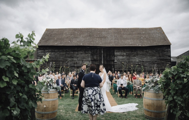 Bride and Groom stand before celebrant in the grounds of an outdoor vineyard wedding with a barn as a back drop