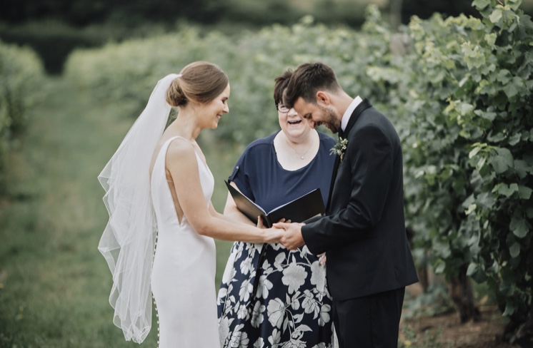 The bride, dressed in white, and groom, dressed in a dinner suit, laugh as they say their vows in front of Shelley the celebrant at their outdoor ceremony at a Vineyard in Essex