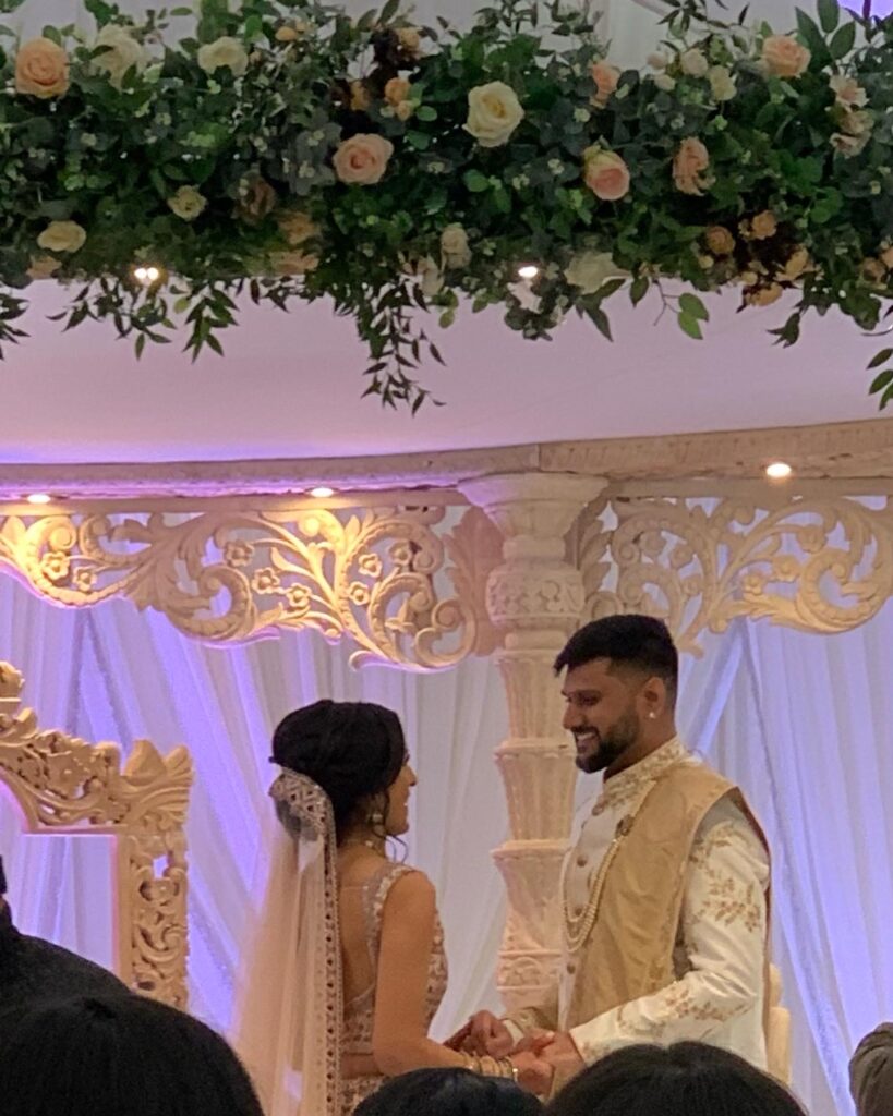 Bride and Groom hold hands and smile at each other under Mandap decorated with flowers