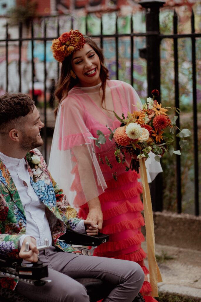 A bride in a bright pink dress carrying a bouquet of flowers holds hands with her husband who is seated in a wheel chair wearing a floral blazer and white shirt.