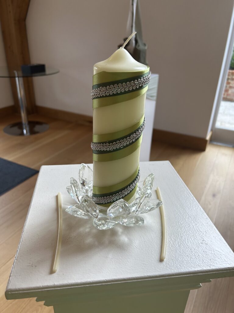 A cream coloured church candle decorated in green and silver ribbon stands in a crystal holder atop a stand.