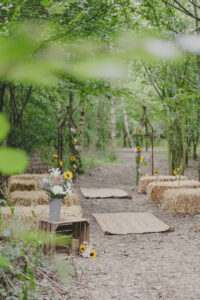 A beautiful woodland enclosure decorated simply with seasonal flowers, hay bale seating and a few rugs to mark the aisle - courtesy of Mannings Farm