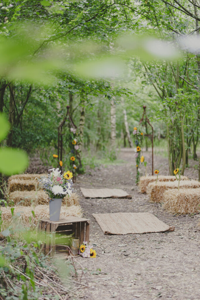 A beautiful woodland enclosure decorated simply with seasonal flowers, hay bale seating and a few rugs to mark the aisle - courtesy of Mannings Farm