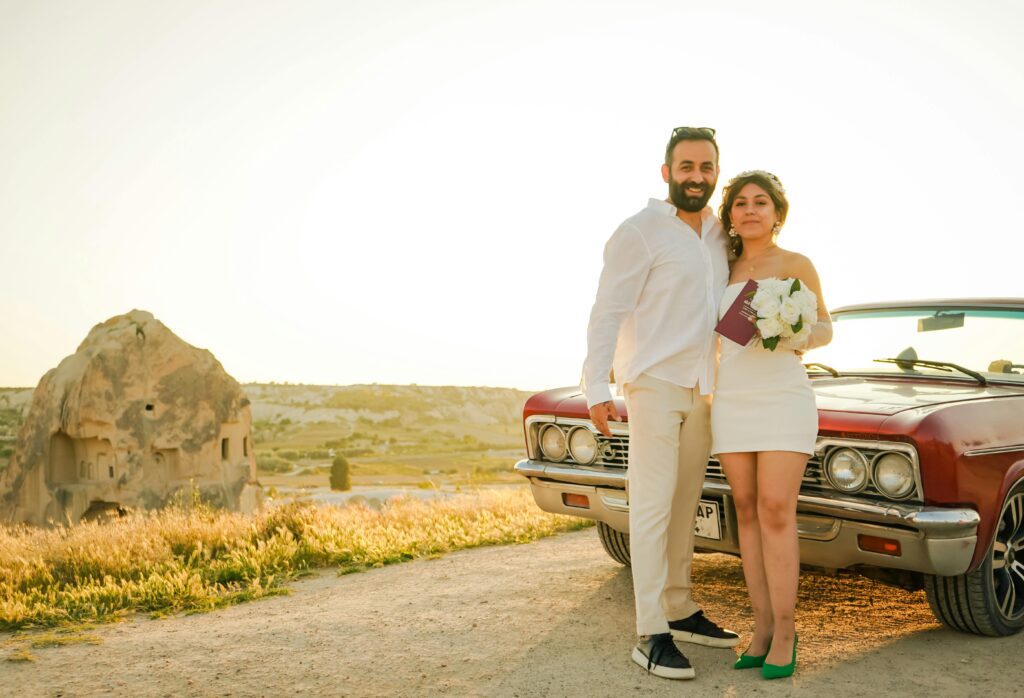 A bearded man in neutral trousers and shirt stands with a woman dressed in white holding a bouquet,  in front of an American muscle car after their vow renewal ceremony