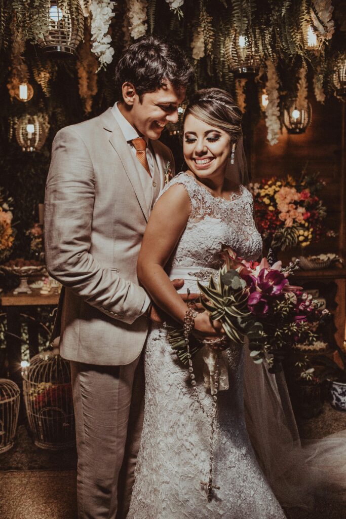 A bride holding a bouquet smiles as her groom clasps her waist infron of a backdrop filled with lights as they renew their vows