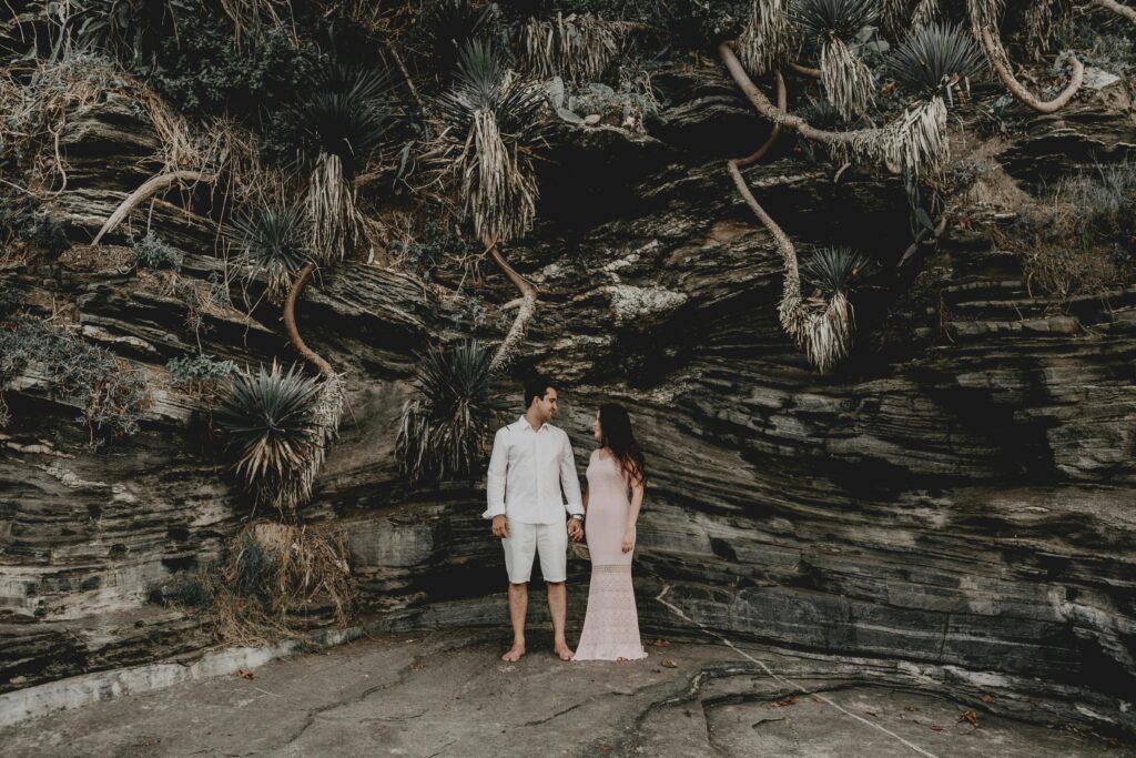 A man in neutral colour shirt and short stands with a woman in a formal dress under a canopy of foliage as they complete their vow renewal ceremony
