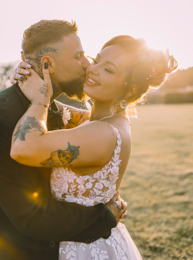 A bride in a white dress and groom in a dark suit, bith with visible tattoos, share a kiss in a field as the sun sets behind them.