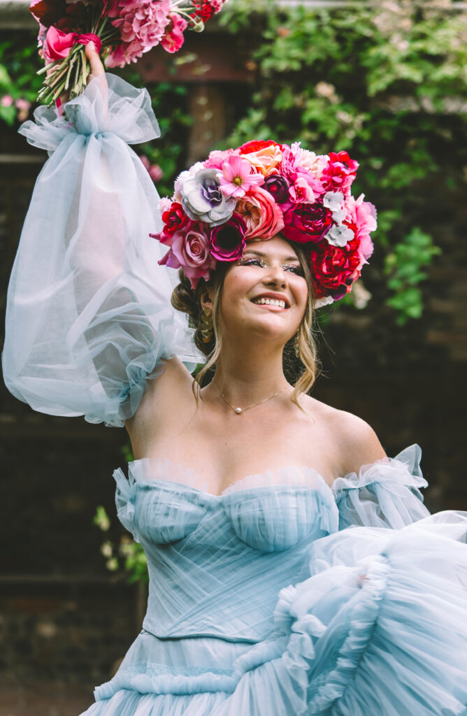 A woman in a blue bridal dress waves a huge bouquet of pink and red flowers above her head and smiles into the distance, respendant in a flower crown of peonies and ranunculas in reds, pinks and oranges.