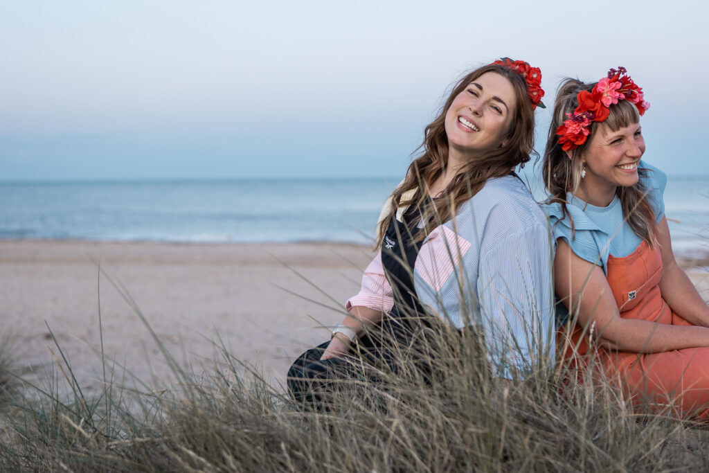Emily and Gemma sit on a sand dune with the sea and setting sun in the background, they are laughing and wearing colourful flower crowns.
