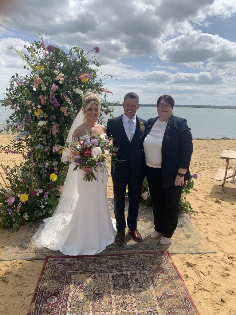 A bride and groom stand alongside their celebrant on a beach in Essex