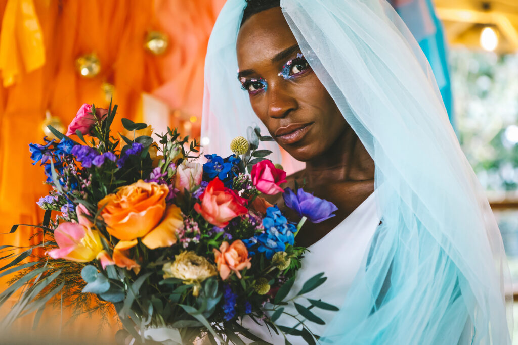 A person of colour wearing a veil looks into the lens of the camera. They are wearing glittering blue eyeshadow with a nude lip colour and holding a bouquest of flowers in orange, pink and blue colurs.