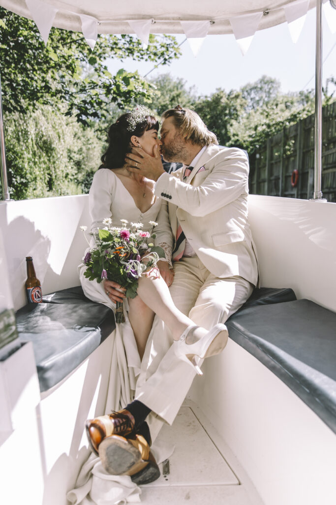 A bride and her groom, both dresses in white sit on a boat and kiss. The bride holds a colourful bouquet of wildflowers.