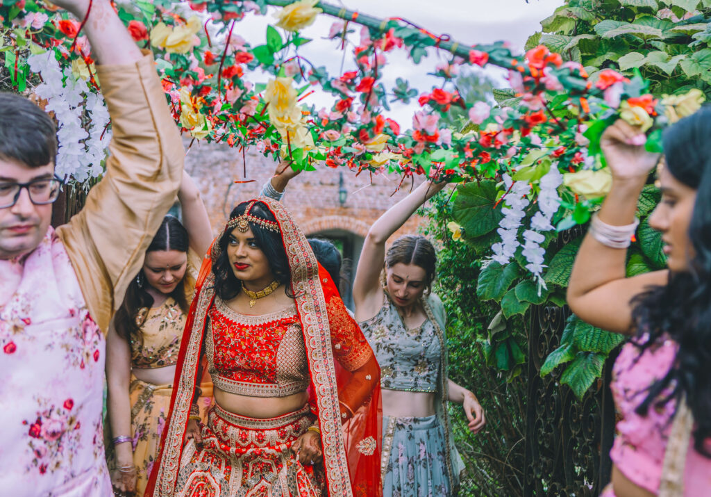 An woman in redand gold Indian dress walks below a structure covered in flower garlands. She is attended by four people in all colourful garments.