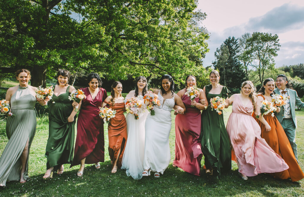 A wedding party in jewel tones dresses and suits form a line of smiles and laughter, with the bride in the centre, in a field with a big oak tree backdrop.