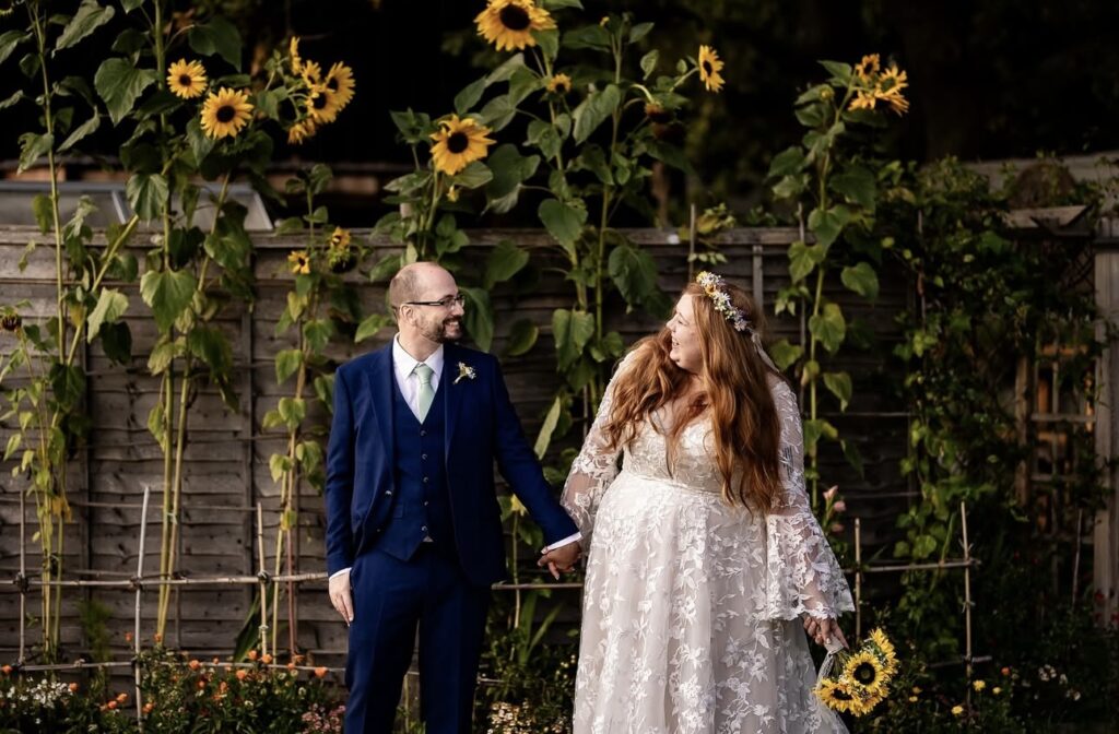A bride and her groom stand hand in hand with sunflowers as the backdrop to the image