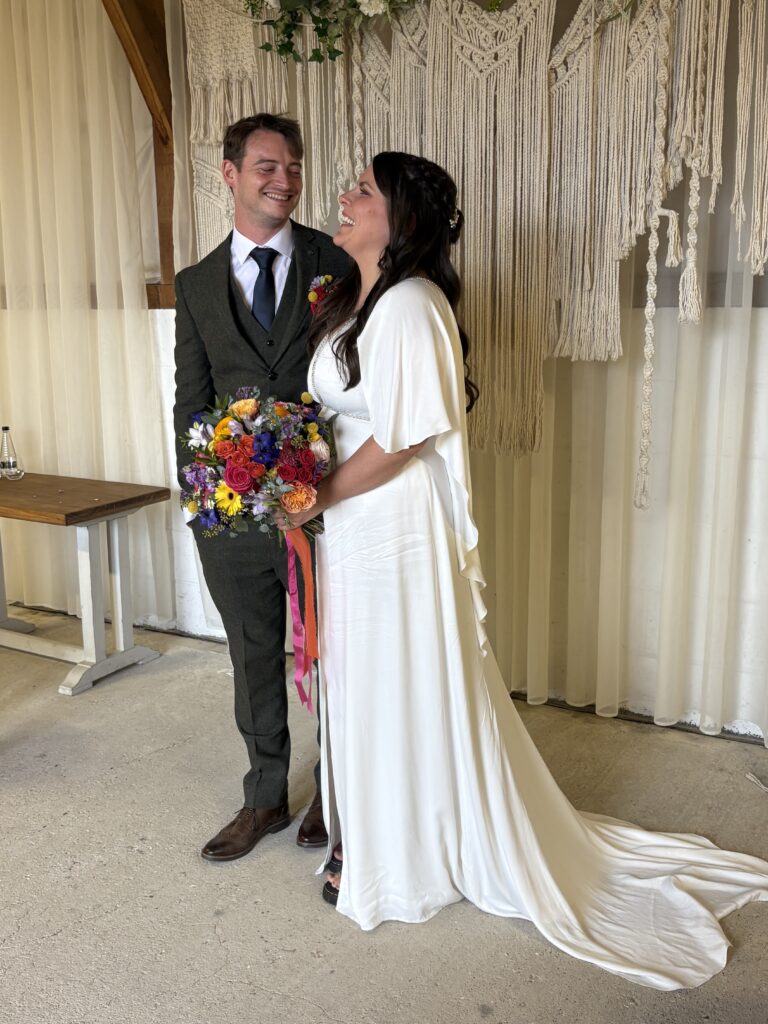 The bride in a white dress holding a colourful bouquet, looks up and smiles at a smiling groom in a green tweed suit.