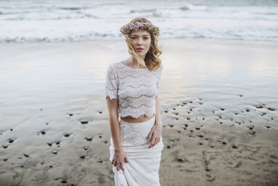 A bride stands on a beach as the tide recedes wearing a flower crown and a two piece white outfit.