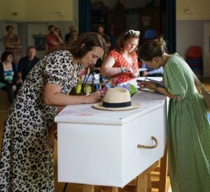 3 ladies lean over a white coffin and write messages on it