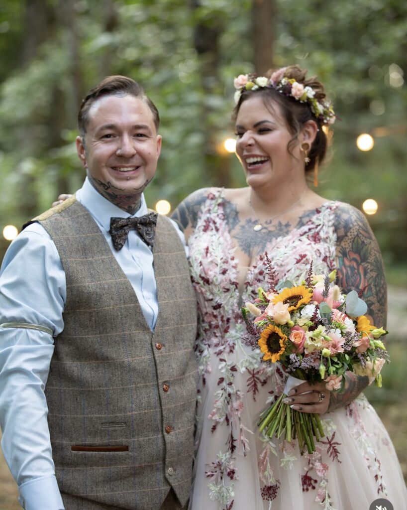 The groom looks into the camera and smiles whilst his bride, holding her bouquet laughs alongside him.