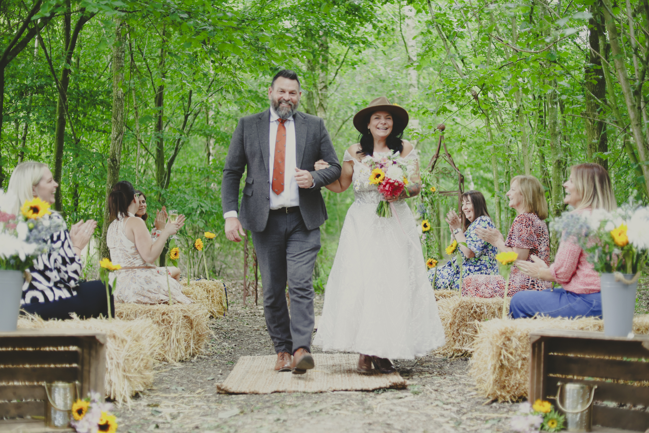The bride in a boho style white dress and sunflower adorned summer hat, walks up the aisle with her groom dresses in a tweed suit after a fantastic ceremony performed by Shelley Bell Celebrant at Mannings Farm